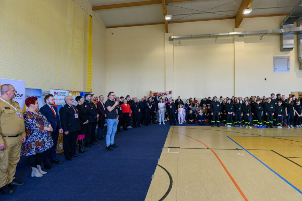 Grupa osób w mundurach stoi w sali gimnastycznej o żółtych ścianach. W tle widoczne plakaty i transparenty. Niebieska wykładzina pokrywa część podłogi.