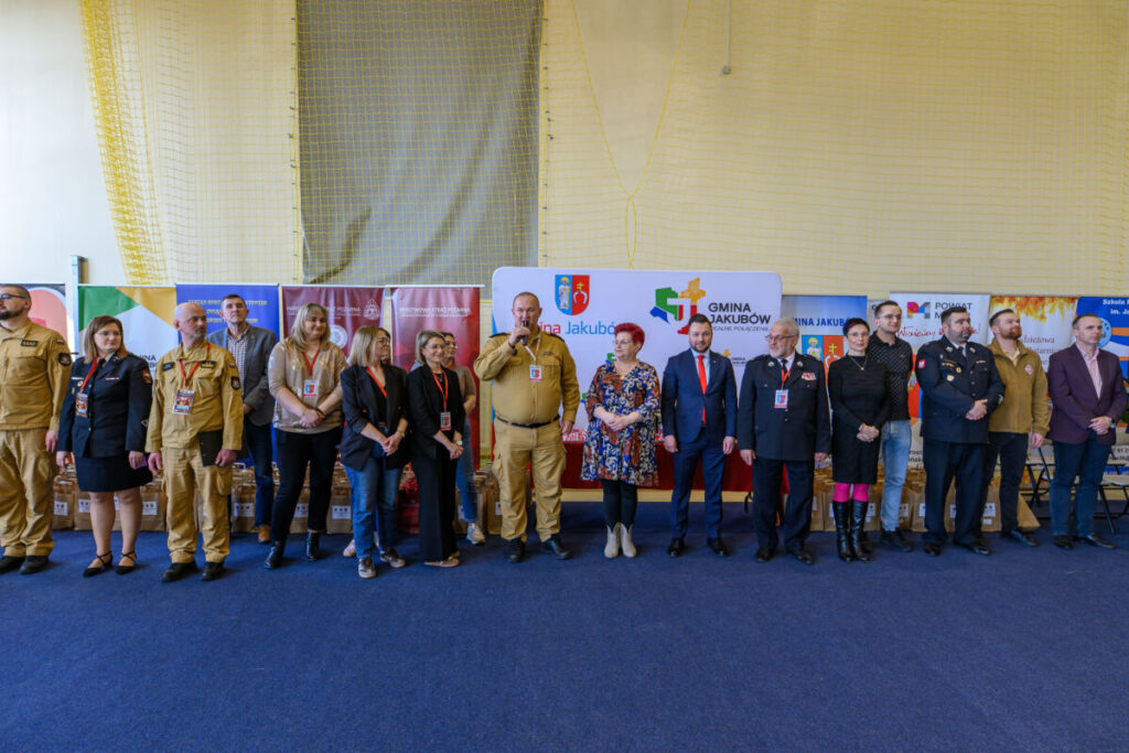 Grupa osób stoi na niebieskiej podłodze, w tle kolorowe bannery i siatka na ścianie. Kobiety i mężczyźni są w różnych strojach, niektórzy w mundurach. Na środku kobieta w kwiecistej sukience, obok mężczyzna