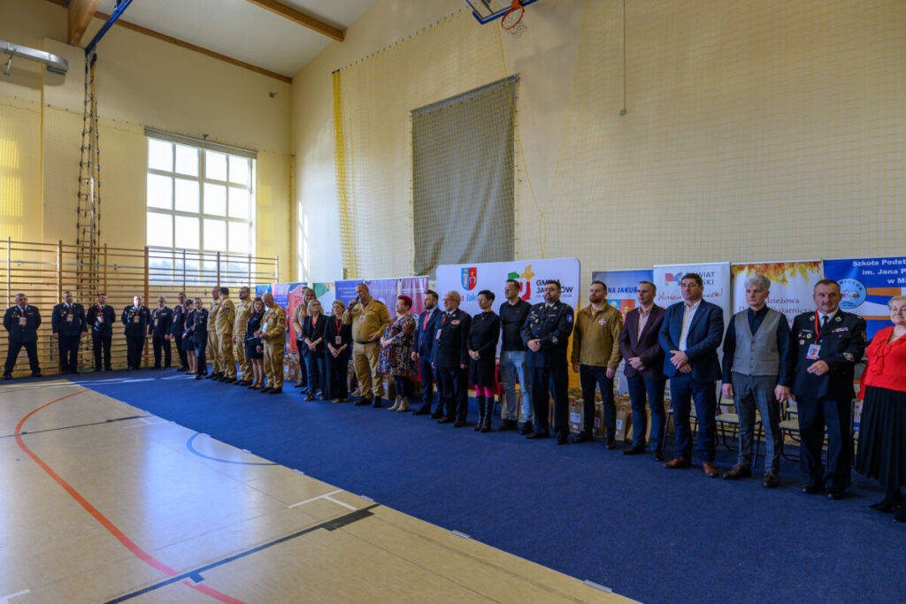 W jasnej sali gimnastycznej stoi grupa ludzi na niebieskim dywanie. Za nimi widać bannery i duże okna. W tle jest jasna ściana z siatką.