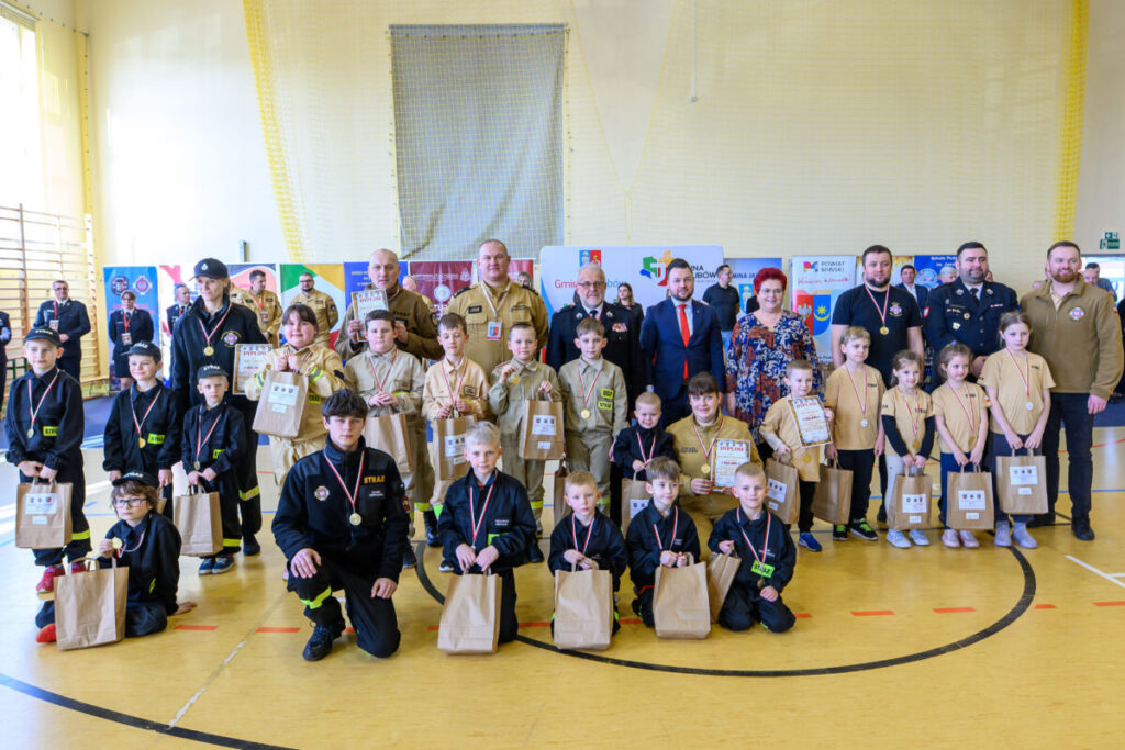 Grupa strażaków i dzieci pozuje z certyfikatami na żółtej sali gimnastycznej. Kolorowe flagi w tle. Wszyscy mają medale.