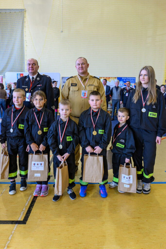 Grupa dzieci w strojach strażackich z medalami na szyjach stoi przed dorosłymi. W tle osoby i banery na jasnym tle. Dzieci trzymają torby papierowe.