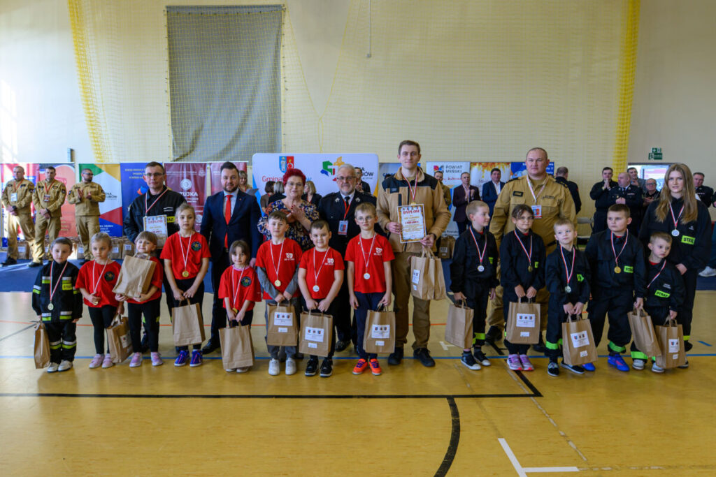 Grupa dzieci i dorosłych w czerwonych i czarnych strojach stoi w sali gimnastycznej. Tło tworzą kolorowe banery. Każdy trzyma torbę i medal.