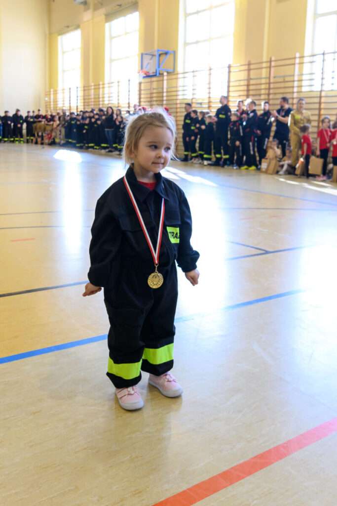 Dziewczynka w mundurze strażackim z medalem stoi na sali gimnastycznej. W tle tłum dzieci w podobnych strojach. Jasne ściany i duże okna.