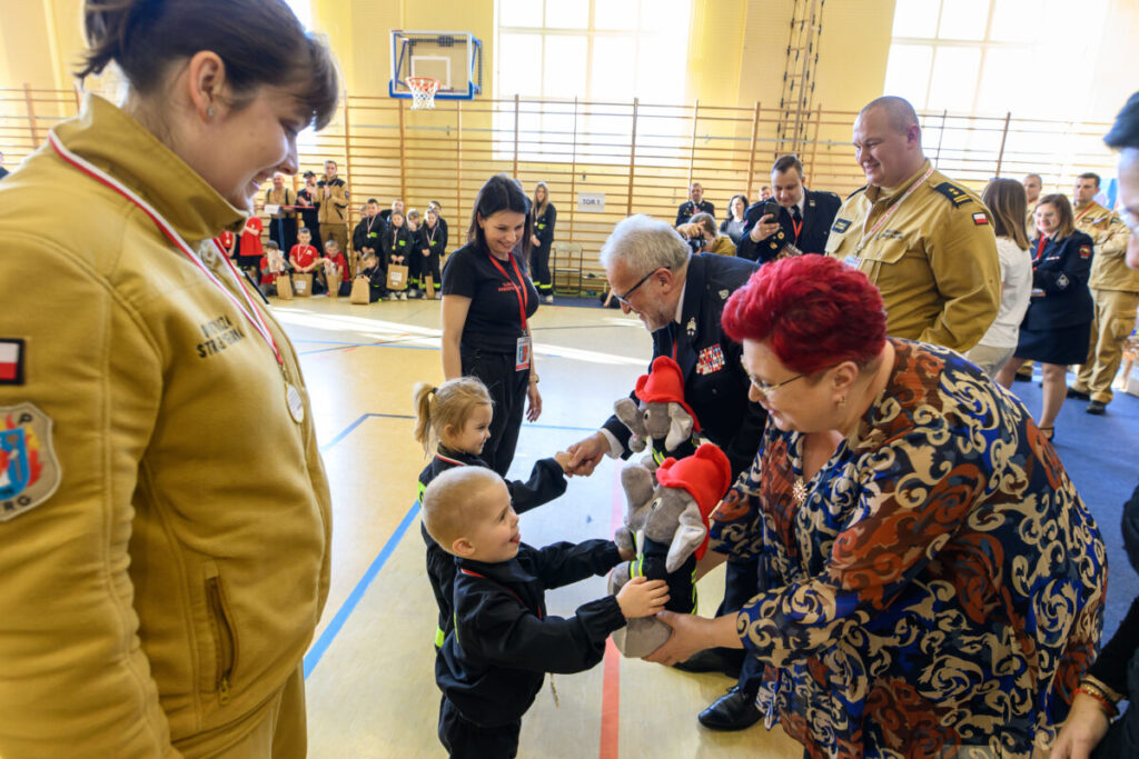 Na zdjęciu widać grupę ludzi w jasnej sali gimnastycznej. Dzieci otrzymują pluszowe maskotki od dorosłych. Ludzie mają na sobie stroje strażackie i formalne.