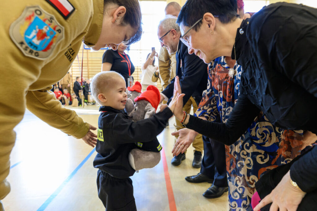 Na zdjęciu dziecko w czarnym stroju z pluszakiem przybija piątkę z dorosłą osobą. Tło jest jasne, wnętrze sali. Obecne są kolorowe stroje.