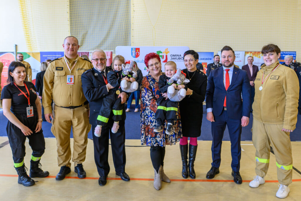 Grupa osób pozuje do zdjęcia w jasnej sali z kolorowymi planszami w tle. Wszyscy są elegancko ubrani, trzymają dwójkę dzieci. Dzieci mają maskotki w kształcie szarych zwierzątek.