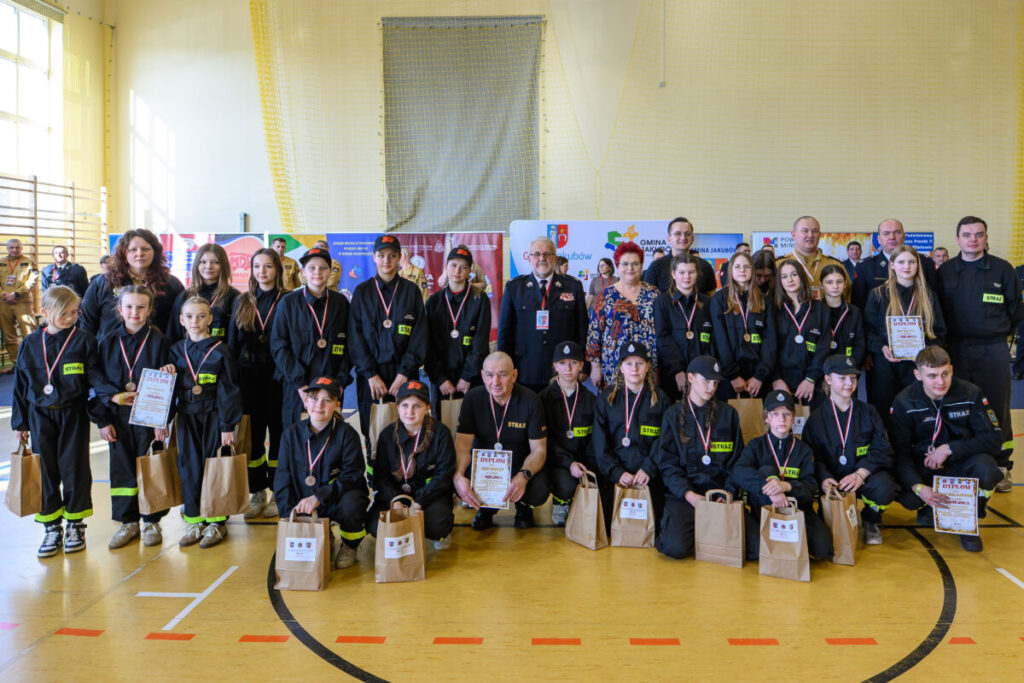 Grupa osób w mundurach strażackich stoi na hali sportowej. Mają medale i dyplomy, trzymają torby. Tło z bannerami promocyjnymi.
