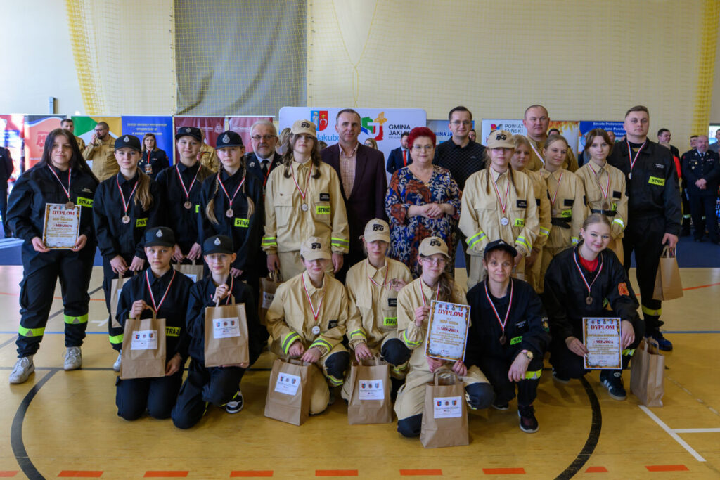 Grupa młodzieży i dorosłych w strojach strażackich stoi na sali sportowej. Jasnożółte i czarne mundury, kilka osób trzyma dyplomy i torby. W tle jasne ściany i banery z napisami.
