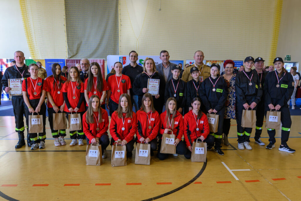 Grupa młodych osób w czerwonych i czarnych mundurach pozuje z medalami. Tło stanowi hala sportowa. Wszyscy trzymają torby papierowe.