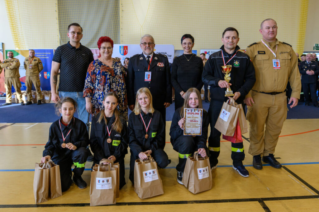 Grupa osób na jasnym tle sali, w mundurach strażackich, z torbami i dyplomem. Kilka osób stoi, inne kucają. W tle banery i obserwatorzy.