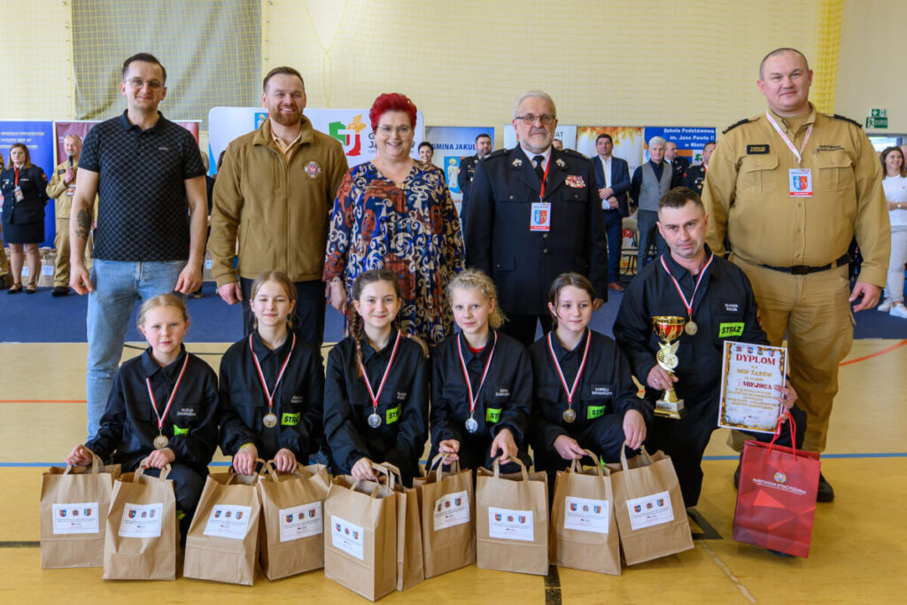 Grupa ludzi z medalami pozuje w jasnej sali, tło z plakatami. Na przodzie dzieci w ciemnych strojach, trzymają nagrody. Dorośli w stoją w dwóch rzędach.