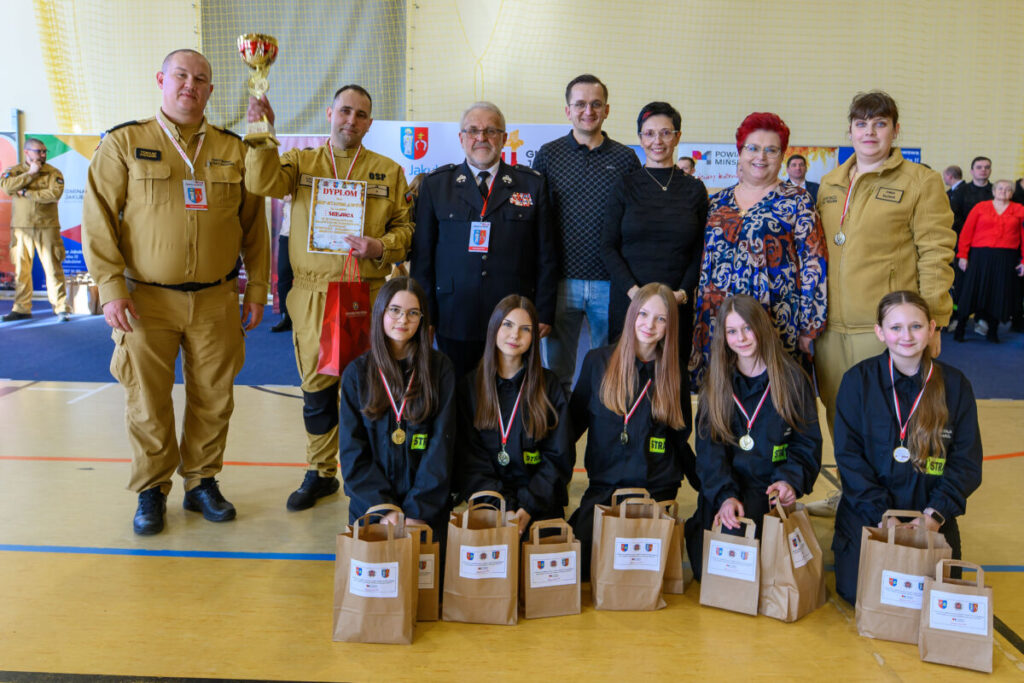 Grupa ludzi pozuje z trofeum i dyplomem. Kobiety na przodzie mają medale i torby. Tło to hala sportowa.