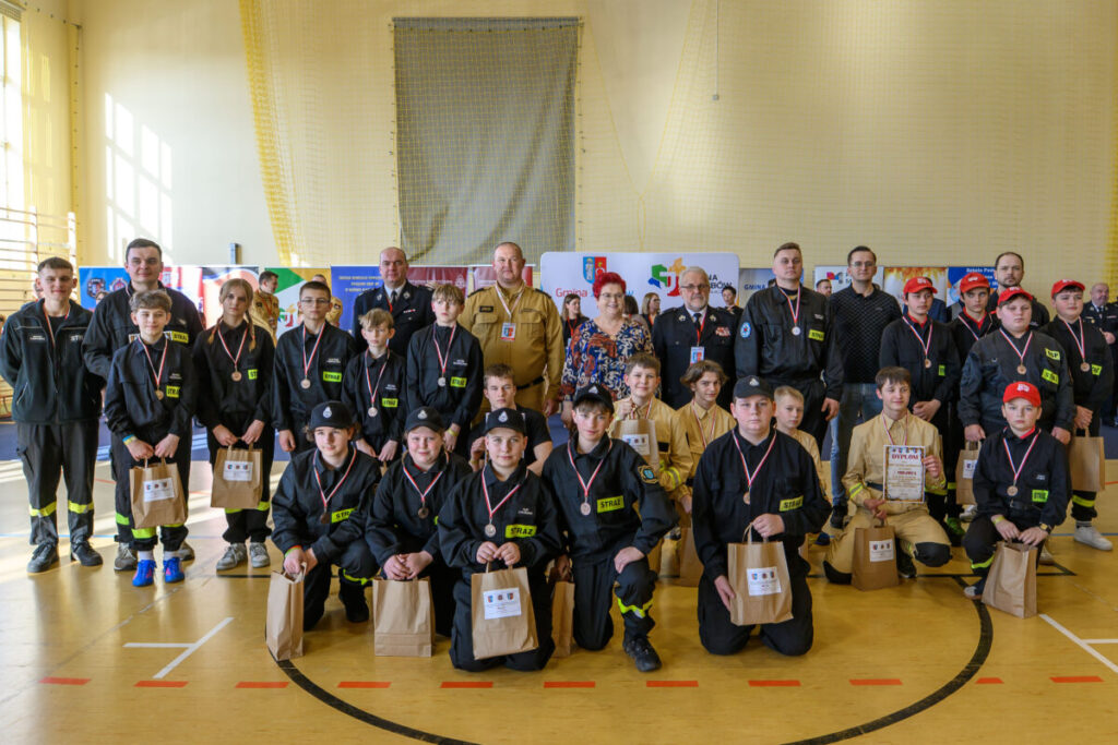 Grupa dzieci i dorosłych w mundurach stoi w sali gimnastycznej, trzymając brązowe torby. Na jasnym tle są kolorowe banery. Uczestnicy noszą medale.