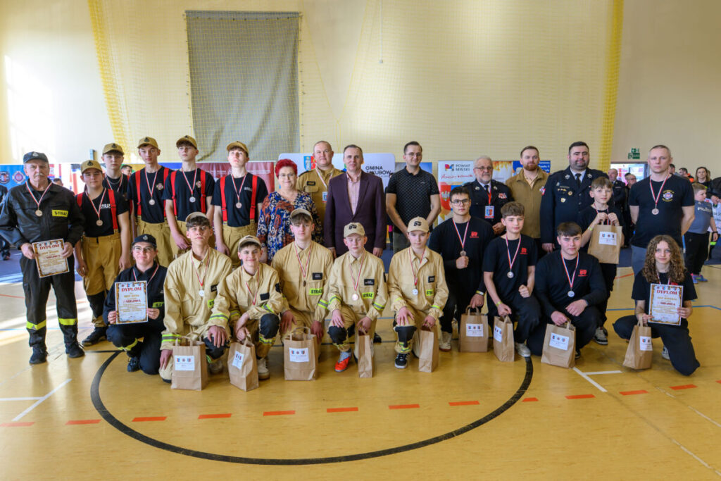 Grupa młodzieży w mundurach stoi w hali, trzyma dyplomy i torby z nagrodami. Dominują kolory czarny i beżowy. Tło jest jasne z siatką ochronną.