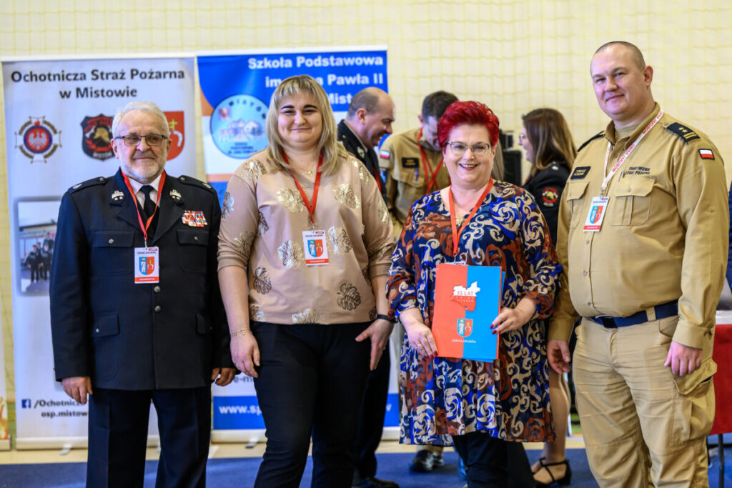 Czworo ludzi stoi przed plakatami w jasnym pomieszczeniu. Dwie osoby noszą mundury strażackie, a jedna trzyma niebieską teczkę. W tle widać banery z napisami.
