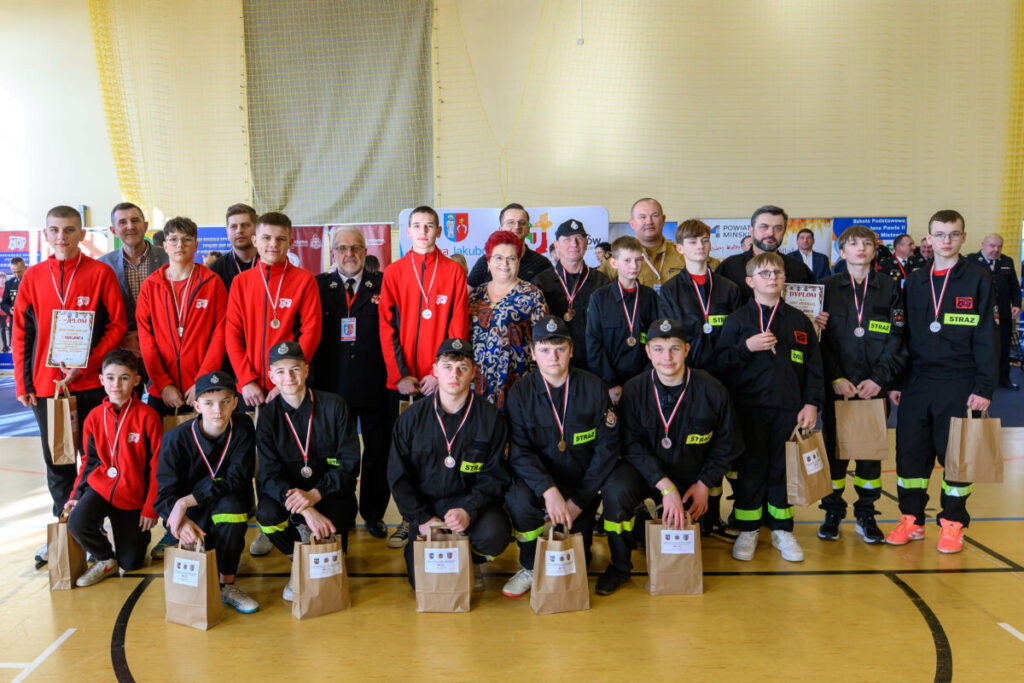 Grupa młodzieży w czarnych i czerwonych strojach strażackich pozuje z medalami. W tle jasne ściany i banery. Na podłodze paczki z nagrodami.
