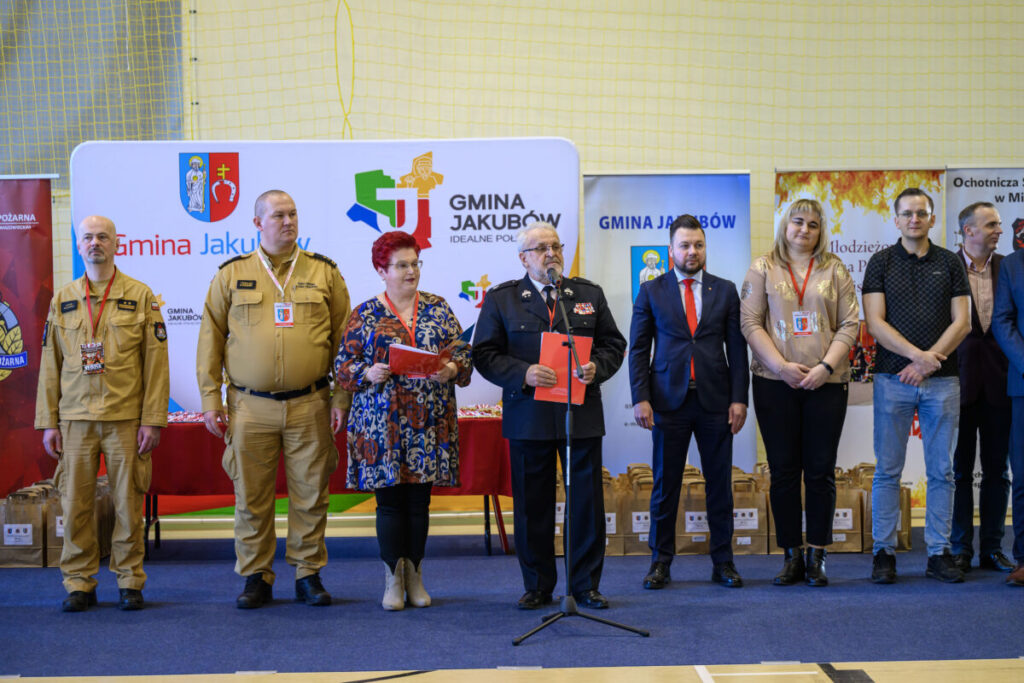 Grupa osób stoi na tle banerów gminnych. Mężczyzna w mundurze trzyma mikrofon. Kolory dominujące to beż, czarny i czerwony.