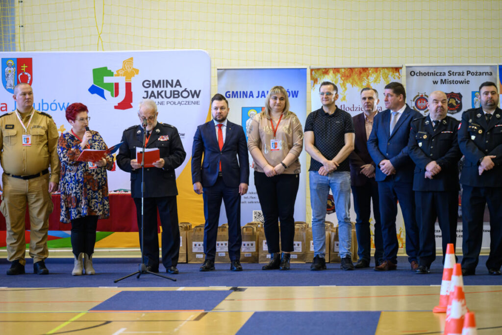 Grupa ludzi stoi na tle banerów z logo gminy Jakubów. Kobieta przemawia do mikrofonu, a mężczyzna czyta z kartki. Kolorystyka jest jasna, z akcentami czerwieni i granatu.