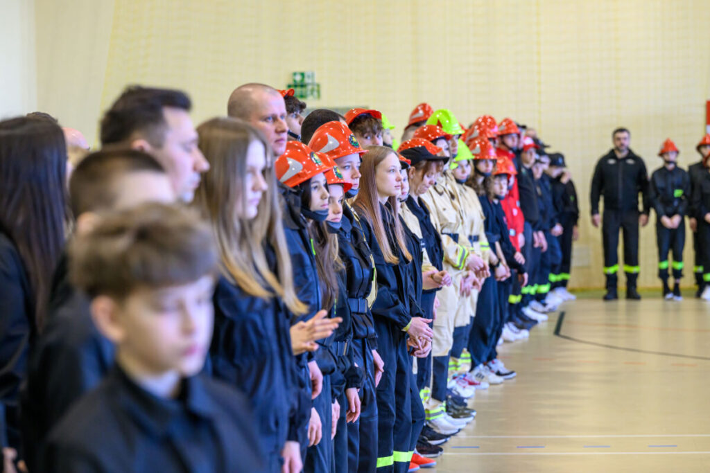 Grupa osób w strażackich uniformach i kaskach w różnych odcieniach czerwieni i beżu stoi w szeregu. Tło jest neutralne, jasne, a cała scena odbywa się w przestronnej hali. Widoczne są detale uniformów i kasków, dodające kolorów.