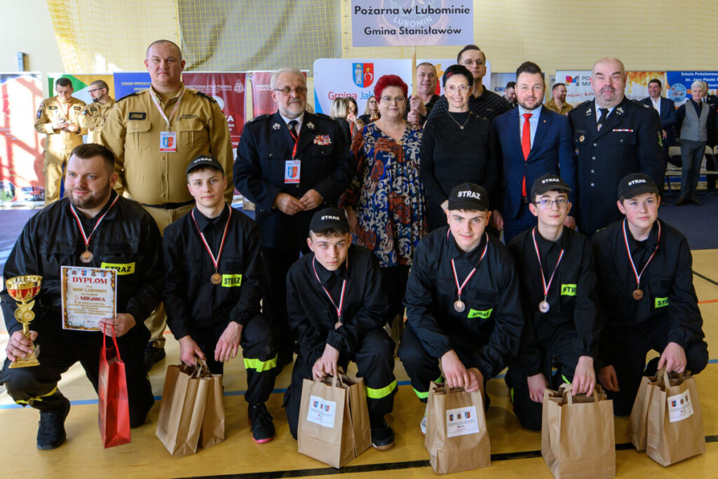 Na zdjęciu grupa strażaków w czarnych uniformach i czapkach, z medalami i torbami. W tle jasna hala, ludzie stojący za nimi. Na tablicy napisy związane z jednostkami strażackimi.