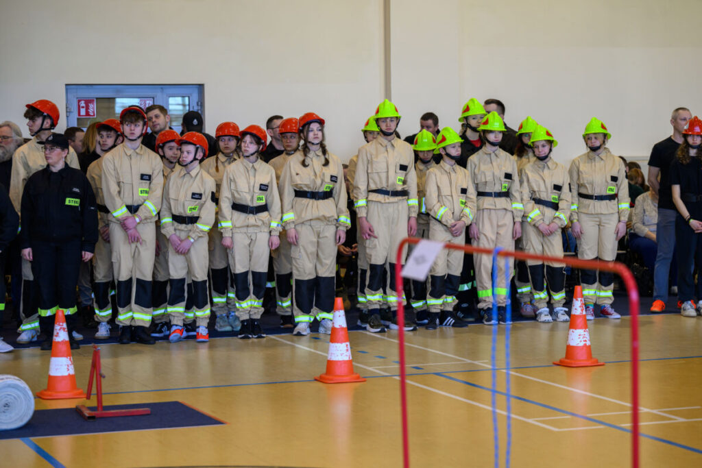 Grupa młodych strażaków w kaskach stoi w sali gimnastycznej. Mają beżowe mundury z odblaskowymi elementami. Przed nimi pomarańczowe pachołki.