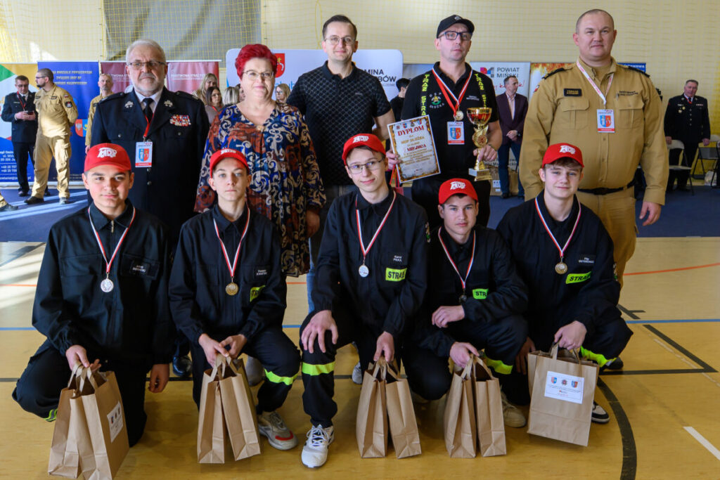 Grupa osób pozuje na sali gimnastycznej. Część jest ubrana na ciemno z czerwonymi czapkami, trzymają torby. W tle banery i inne osoby.