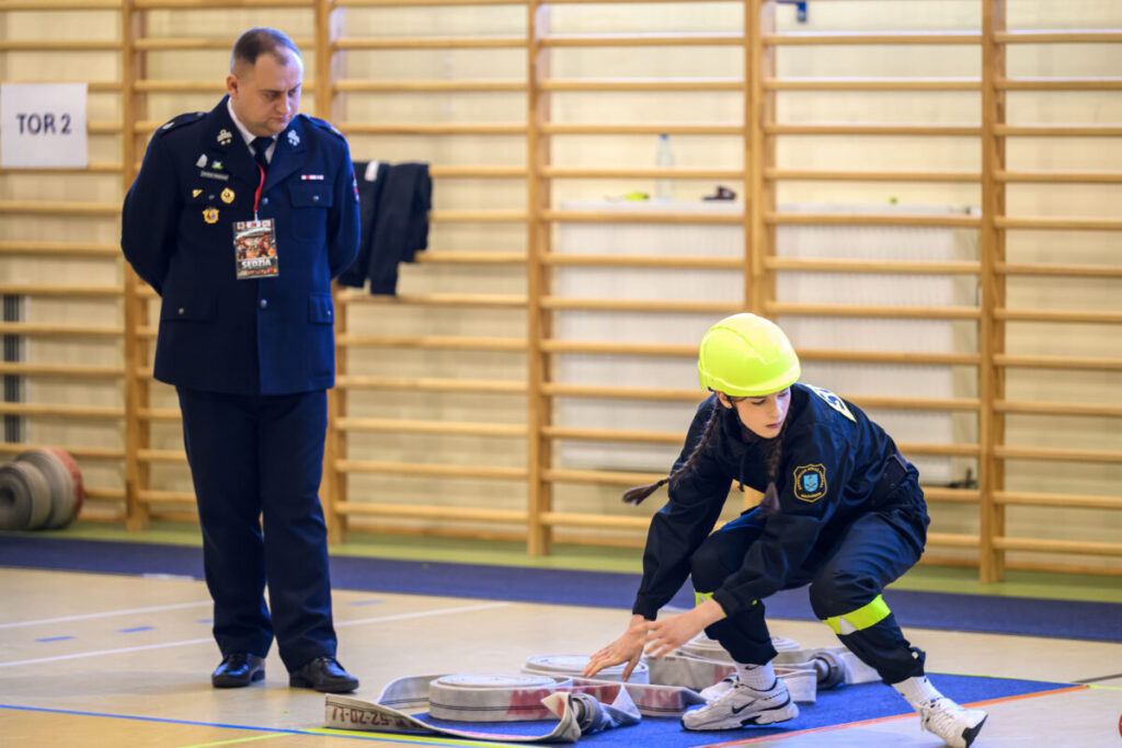 Na zdjęciu młoda osoba w kasku układa wąż strażacki, obok stoi sędzia w mundurze. Tło stanowi drewniana drabinka. Kolory są jasne, dominują niebieski i żółty.