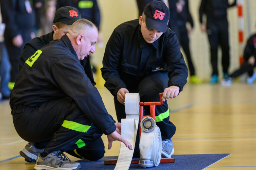 Na zdjęciu dwóch strażaków w czarnych mundurach przygotowuje sprzęt. Jeden z nich klęczy i trzyma wąż strażacki na stojaku. W tle osoby w podobnych strojach na sali gimnastycznej.