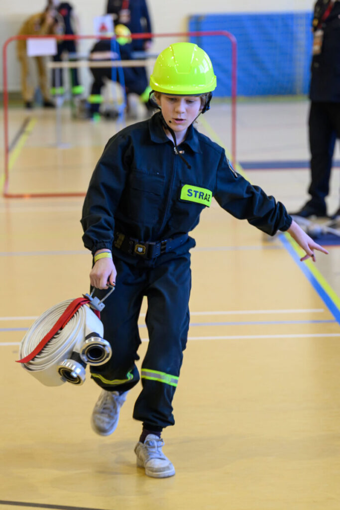 Młody strażak z hełmem niesie wąż strażacki. Tło to sala gimnastyczna z niebieską matą. Stroje są czarne z zielonymi elementami.