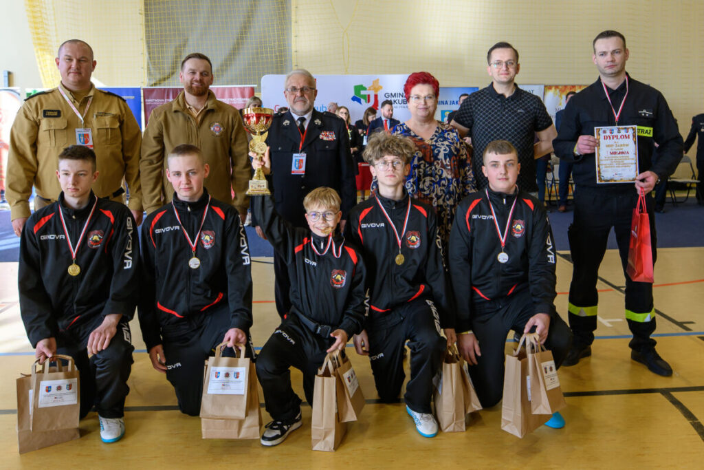 Grupa osób w sportowych strojach z nagrodami stoi na sali gimnastycznej. Trzymają medale, puchar i dyplom. Tło jest jasne z siatką oraz banerami reklamowymi.