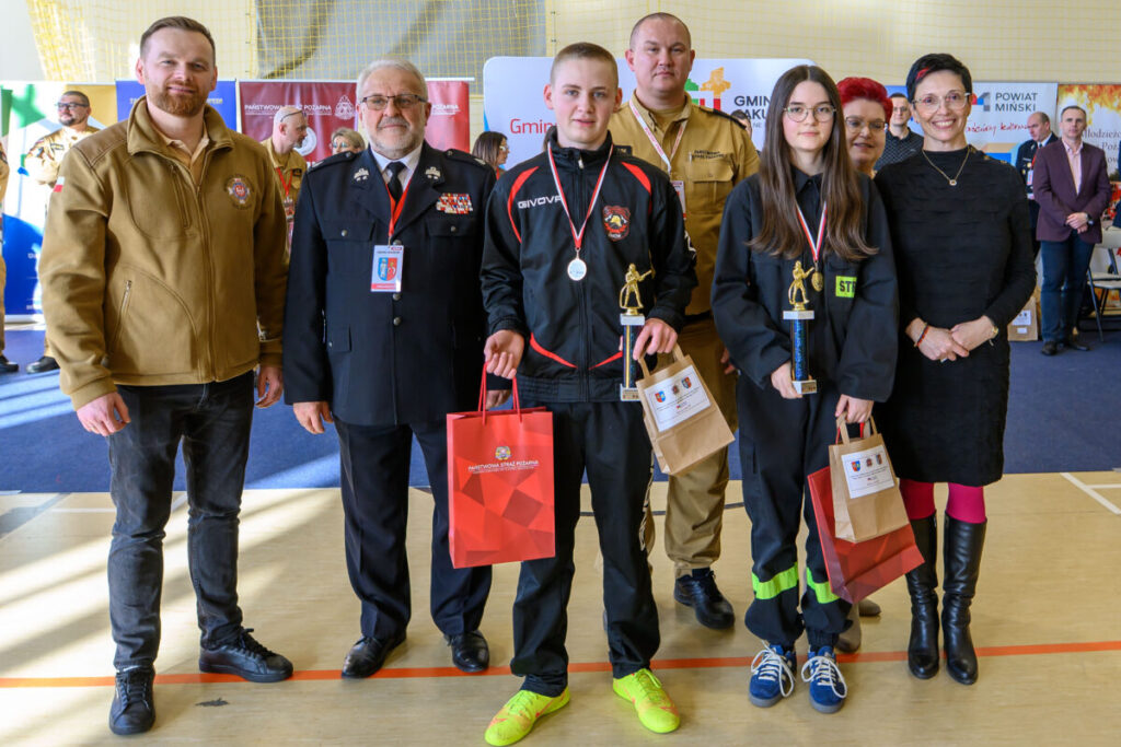 Grupa osób stoi na hali sportowej. W centrum młodzi ludzie z pucharami i medalami. Tło kolorowe z banerami promocyjnymi.