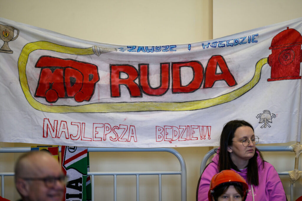 Baner z napisem MDP RUDA, żółtym wężem i czerwonym hydrantem, na białym tle. Tło kremowe, pod banerem siedzą ludzie.