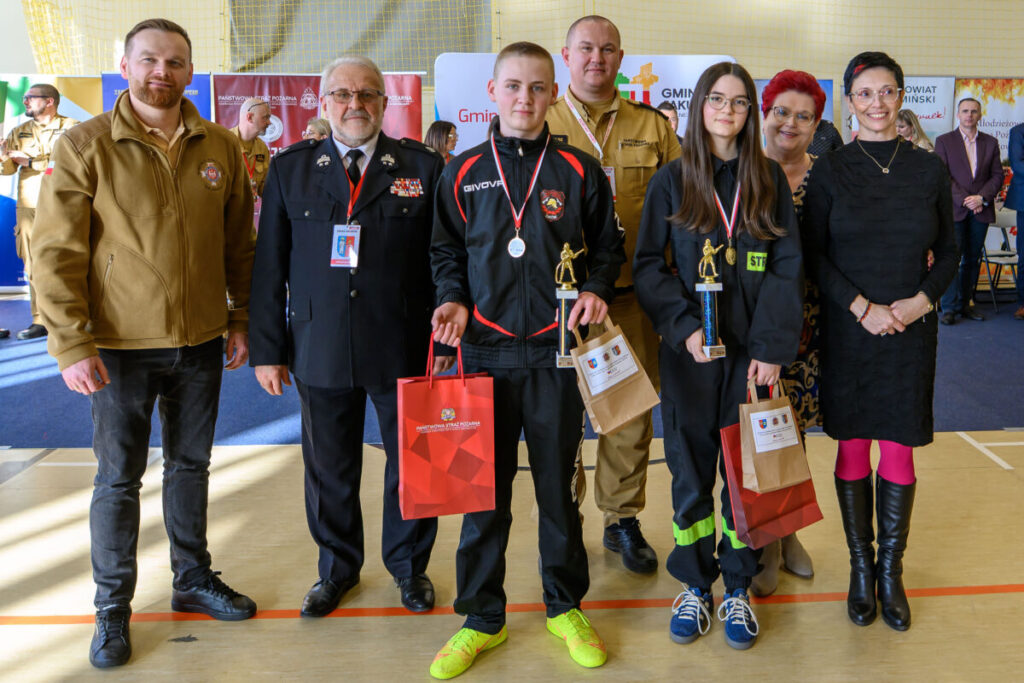 Grupa ludzi z nagrodami na zawodach, kolorowe tło z banerami. Osoby noszą stroje strażackie i garnitury. Przyjazna atmosfera.