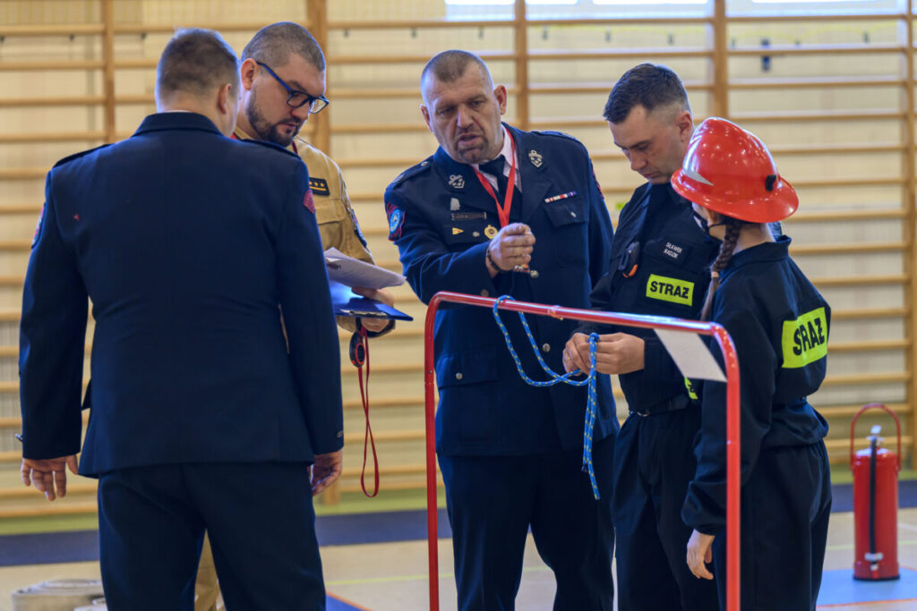 Na zdjęciu widocznych jest pięciu strażaków omawiających zadanie w jasnym pomieszczeniu. Dwóch z nich trzyma linę, jedna osoba w czerwonym hełmie. W tle drewniane elementy do ćwiczeń.