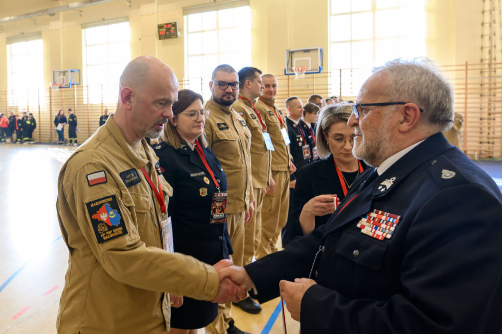 Grupa osób w mundurach stoi w sali gimnastycznej, wymieniając uściski dłoni. Jasne, naturalne oświetlenie. W tle drabinki gimnastyczne.