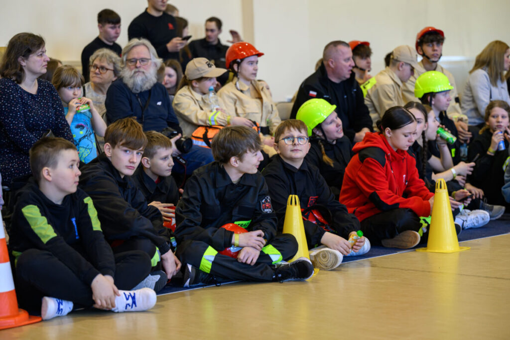 Grupa młodych ludzi siedzi na podłodze w środku sali, ubrani w ciemne ubrania i kolorowe kaski. Na pierwszym planie żółte pachołki. W tle inne osoby w beżowych uniformach.