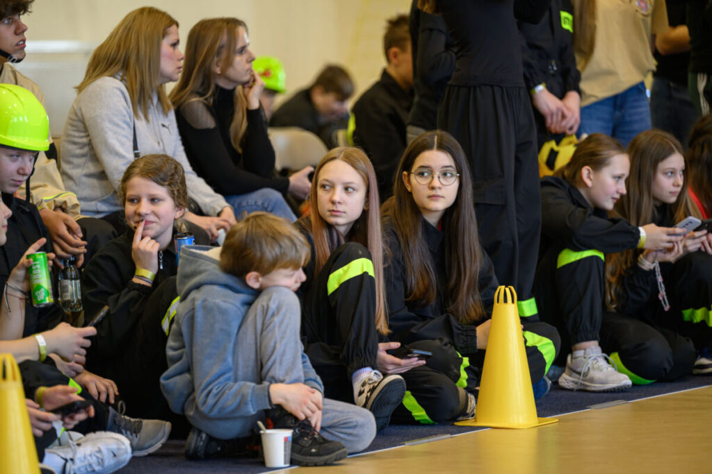 Grupa młodych osób siedzi na podłodze, patrząc na coś poza kadrem. Noszą czarne ubrania z zielonymi akcentami. W tle widoczne jasne ściany i kilka żółtych pachołków.