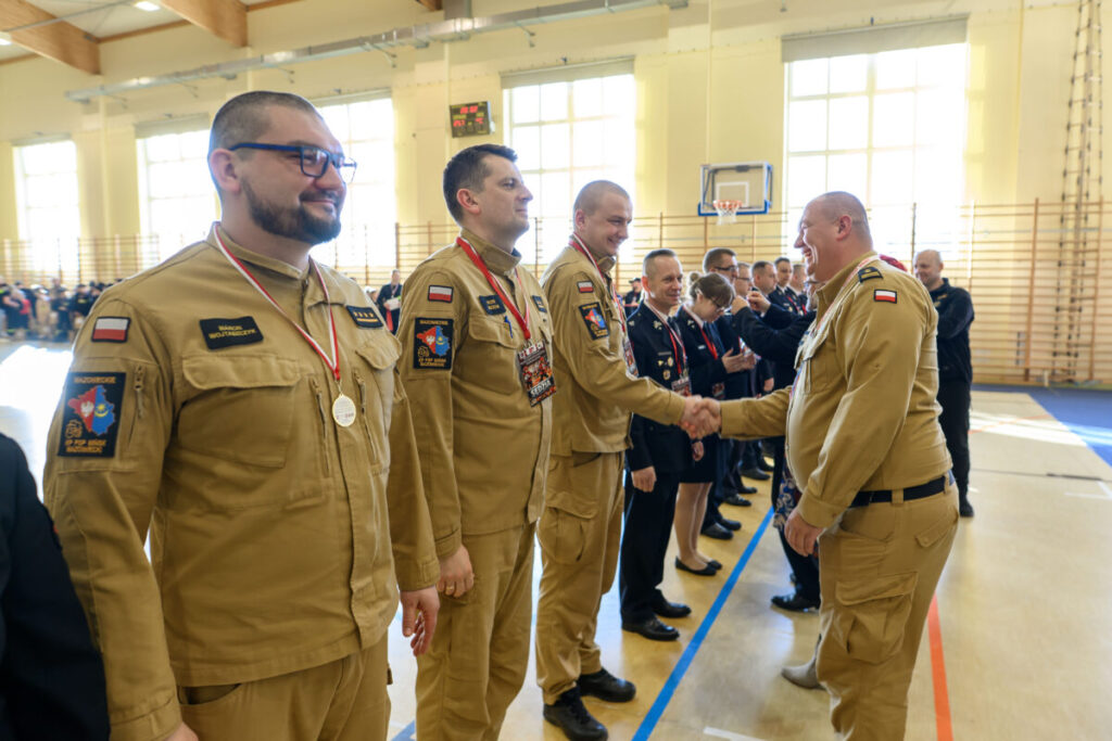 Grupa ludzi w beżowych mundurach w sali gimnastycznej, odbiera medale. Tło to jasne ściany z oknami. Uroczystość z gratulacjami.