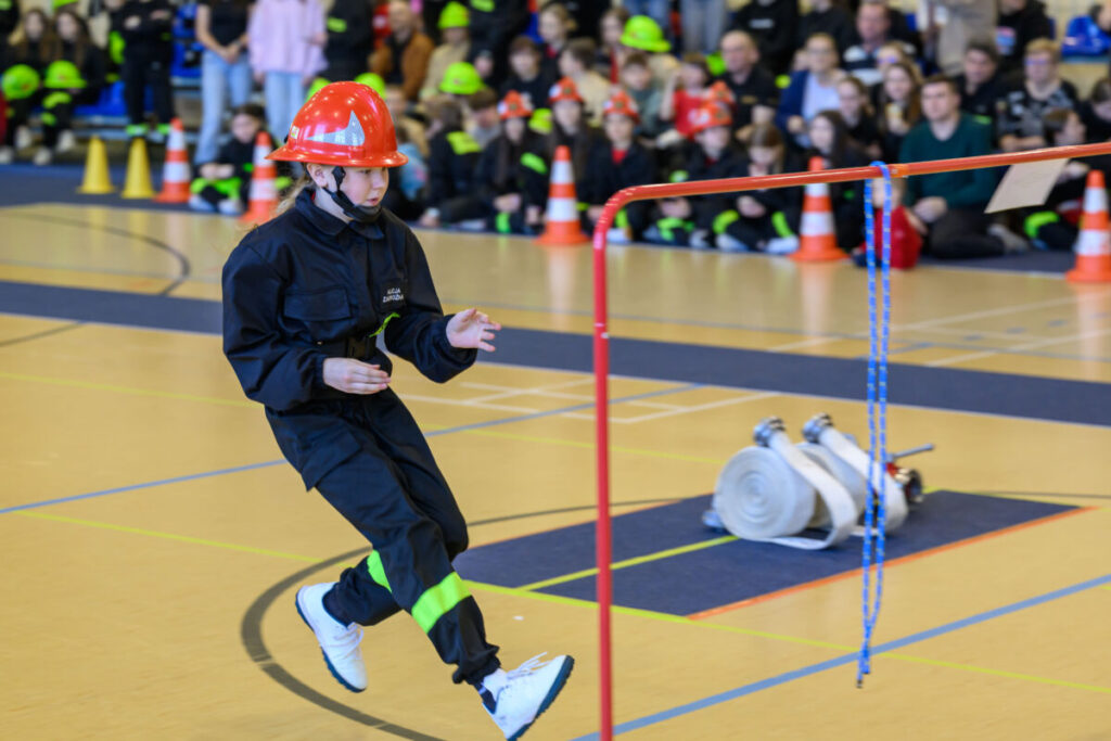 Młoda osoba w czerwonym kasku i czarnym stroju strażackim biegnie po sali gimnastycznej. W tle siedzi grupa dzieci w kolorowych kaskach. Obok kolorowe pachołki i zwinięty wąż strażacki.