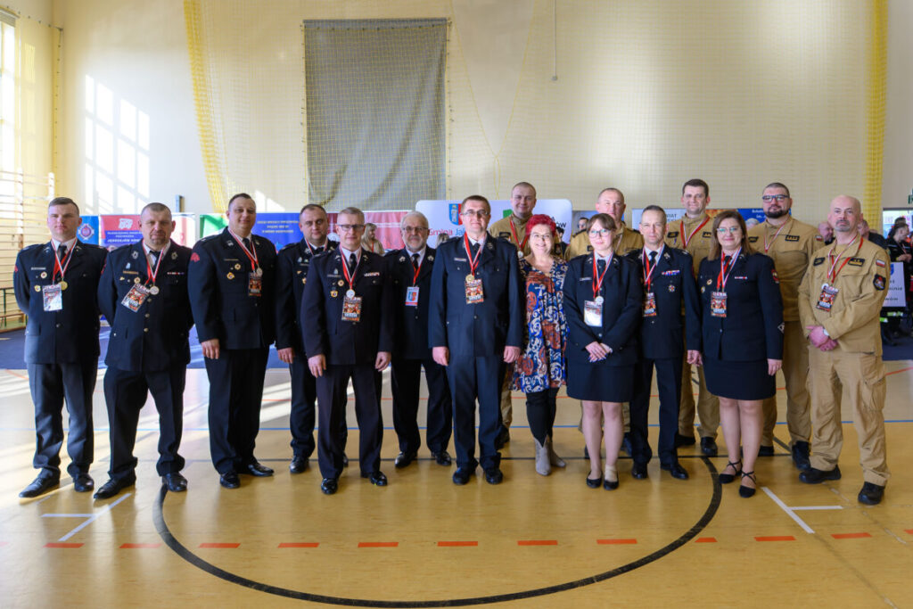 Grupa ludzi w mundurach stoi w świetle hali gimnastycznej. Na ścianie tło z flagami i banerami. Podłoga jasna z liniami.