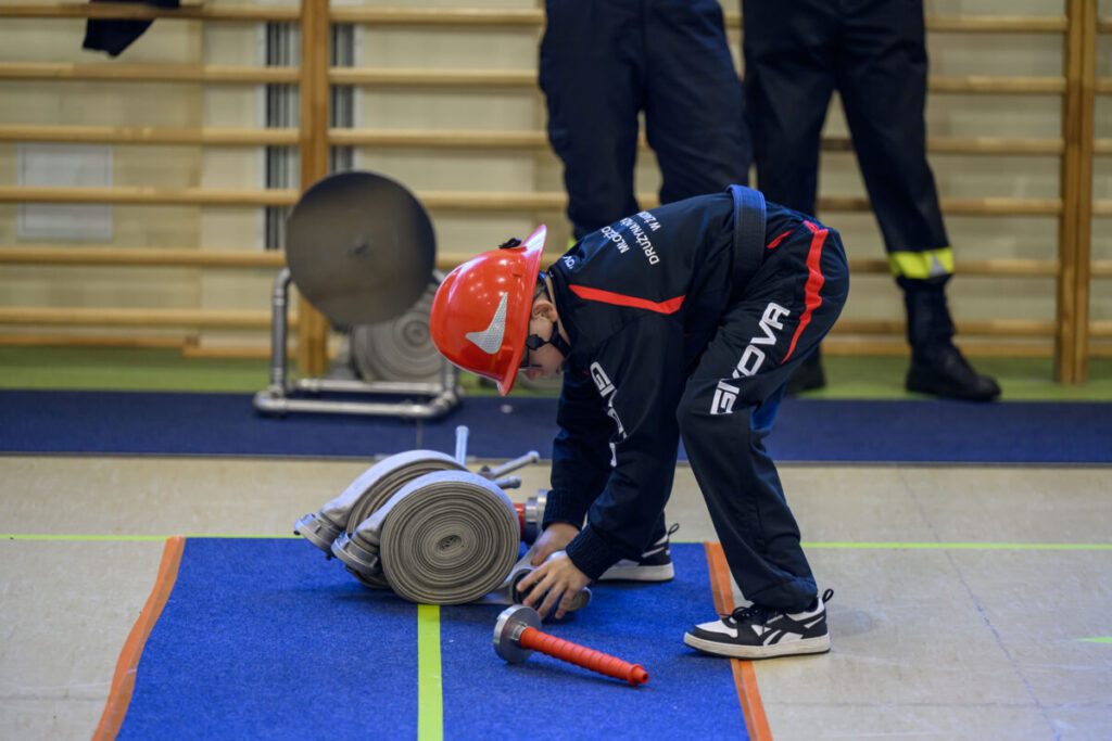 Dziecko w czerwonym kasku ćwiczy z wężem strażackim na niebieskiej macie. Jasne wnętrze z drabinką w tle. Obok leży czerwona końcówka węża.
