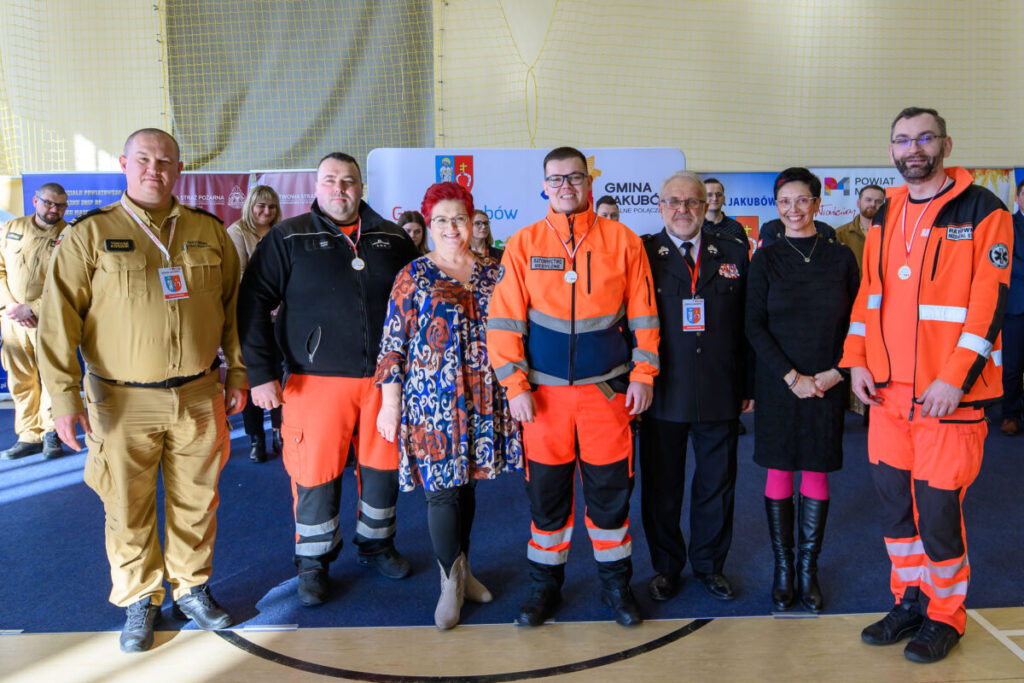 Grupa osób w strojach ratowników i cywilnych pozuje na tle żółtej ściany. Ubrania w kolorach pomarańczowym, beżowym i czarnym. W tle widoczne są banery informacyjne.