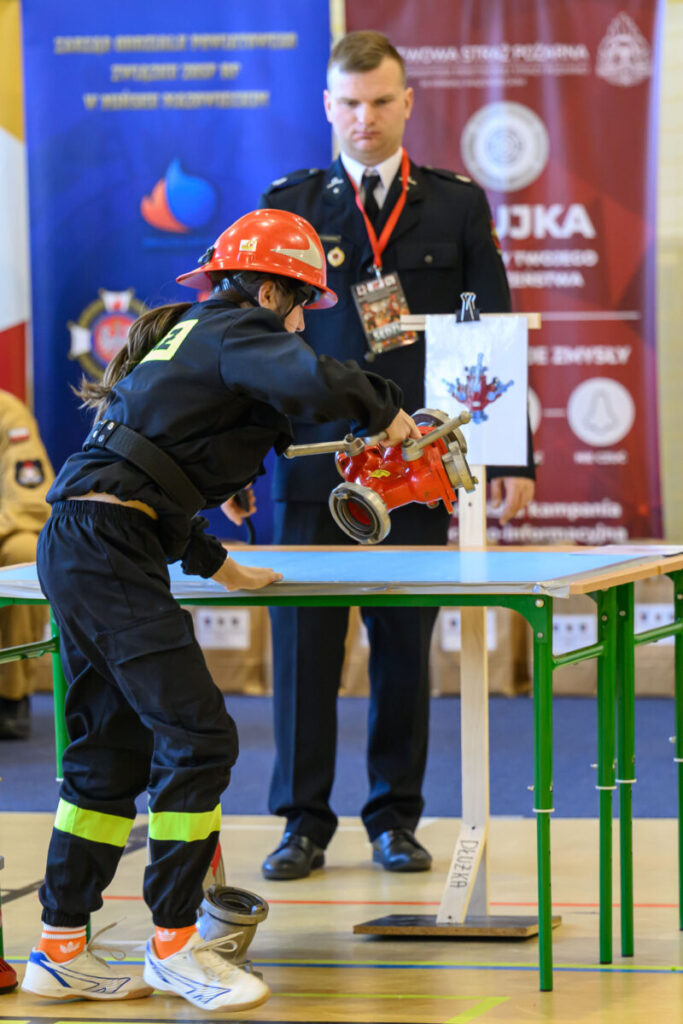 Na zdjęciu młoda osoba w stroju strażaka obsługuje sprzęt na stole. W tle stoi dorosła osoba w mundurze, z niebieskim i czerwonym banerem. Stoły są zielone, a kask osoby jest czerwony.