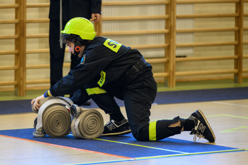Osoba w stroju strażaka i neonowym kasku układa wąż strażacki na podłodze. Tło to drewniana drabinka gimnastyczna. Kolory dominujące to czarny, zielony i beżowy.