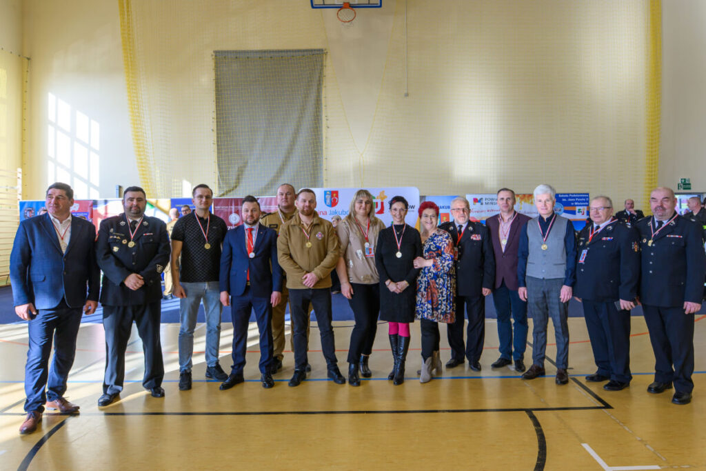 Grupa osób stoi w jasnej sali gimnastycznej, niektórzy w mundurach, z medalami na szyi. Tło zawiera banery z logo. Dominują kolory jasny beż i żółty.