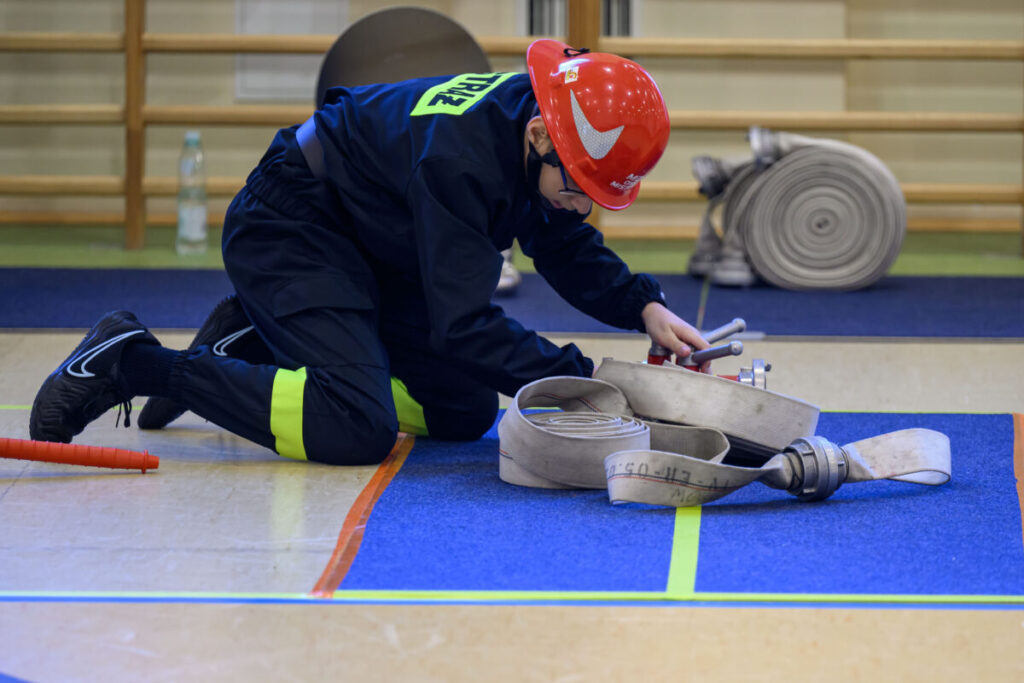 Młody strażak w czerwonym kasku klęczy na niebieskiej macie, układając węże strażackie. Na nogach ma czarne buty i strój z żółtym akcentem. W tle widać drewnianą drabinkę i zwinięty