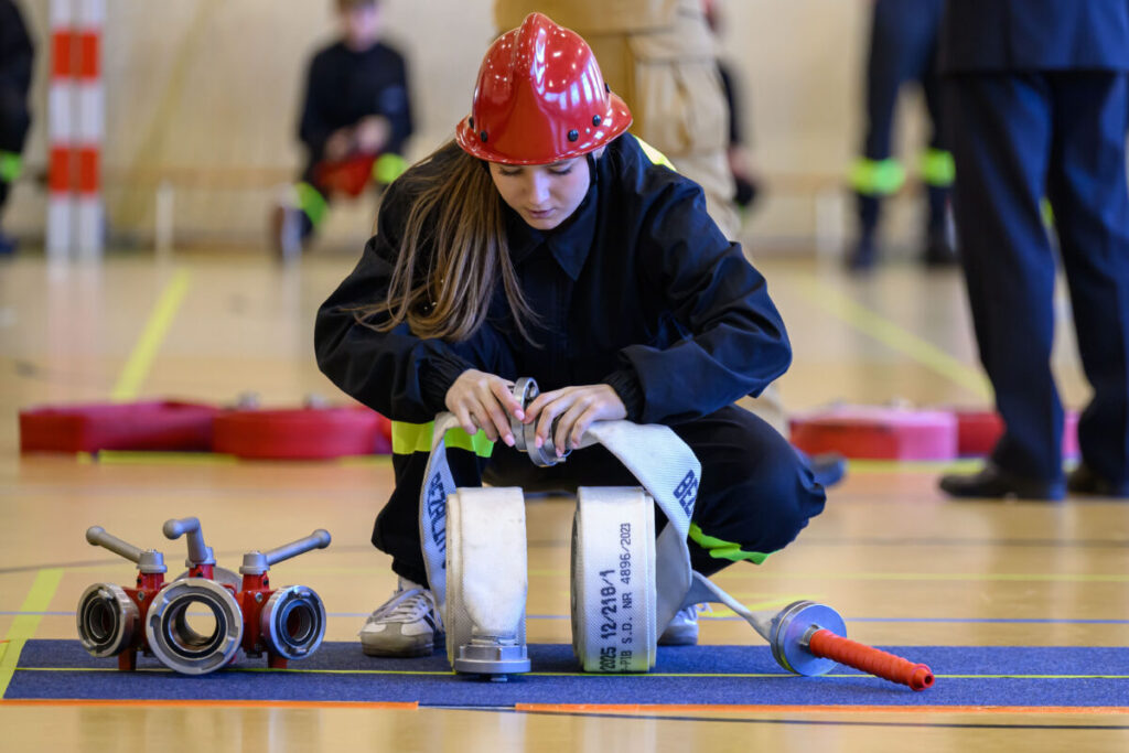 Na zdjęciu młoda osoba w czerwonym kasku układa wąż strażacki na siłowni. Ubrana w czarny strój z odblaskami. Tło jest rozmyte, z widocznymi innymi uczestnikami.