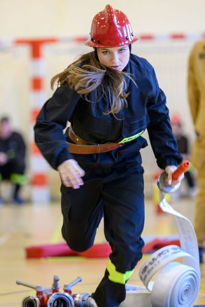 Młoda kobieta w czerwonym hełmie strażackim i czarnym stroju biegnie z wężem po hali. Jasne tło, rozmyta publiczność. Wygląda na skupioną.