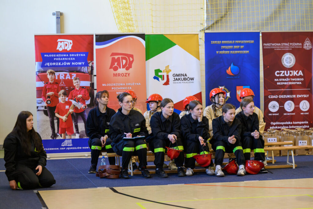 Grupa młodzieży w straży pożarnej, ubrana w czarne mundury i czerwone kaski, siedzi przed kolorowymi banerami na tle sali.