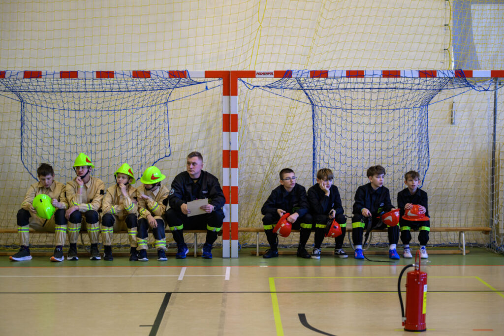 Grupa młodych osób w kaskach siedzi na ławce w hali sportowej. Tło z siatki i bramki, dominują kolory czerwony i żółty. Na podłodze stoi czerwony gaśnica.